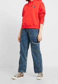 Red hooded sweatshirt with front kangaroo pocket, paired with blue denim cargo pants featuring side pockets. Worn with leopard print sneakers.