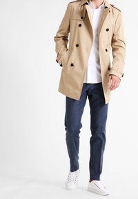 Beżowy trench coat z podwójnym zapięciem, czarnymi guzikami i naramiennikami. Połączony z granatowymi spodniami i bia�łymi trampkami.