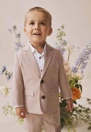 Ung dreng i beige jakkesæt og hvid skjorte står smilende med blomsterarrangement i blødt fokus i baggrunden.