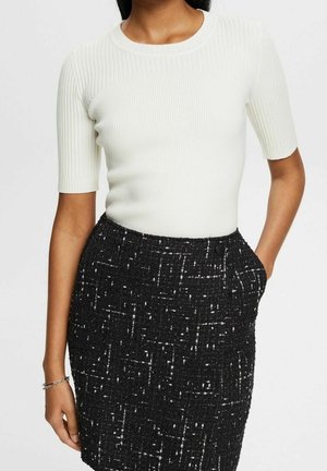 Vrouw draagt een wit geribd topje met korte mouwen, ingestopt in een hoge taille zwarte tweed rok met witte spikkels, handen ontspannen langs haar zij.