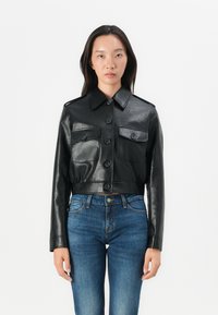 Kuttet svart skinnjakke med to frontlommer, knappestenger og spiss krage, iført over blå denimjeans.