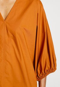 Gros plan d'une personne portant un haut orange avec un décolleté en V profond et des manches trois-quarts amples et froncées.