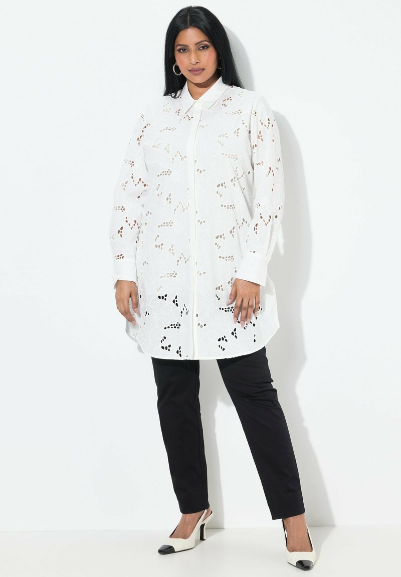 Blusa bianca a maniche lunghe con design floreale a laser cut, chiusura frontale con bottoni e orlo arrotondato, abbinata a pantaloni neri aderenti e scarpe con tacco bicolore.