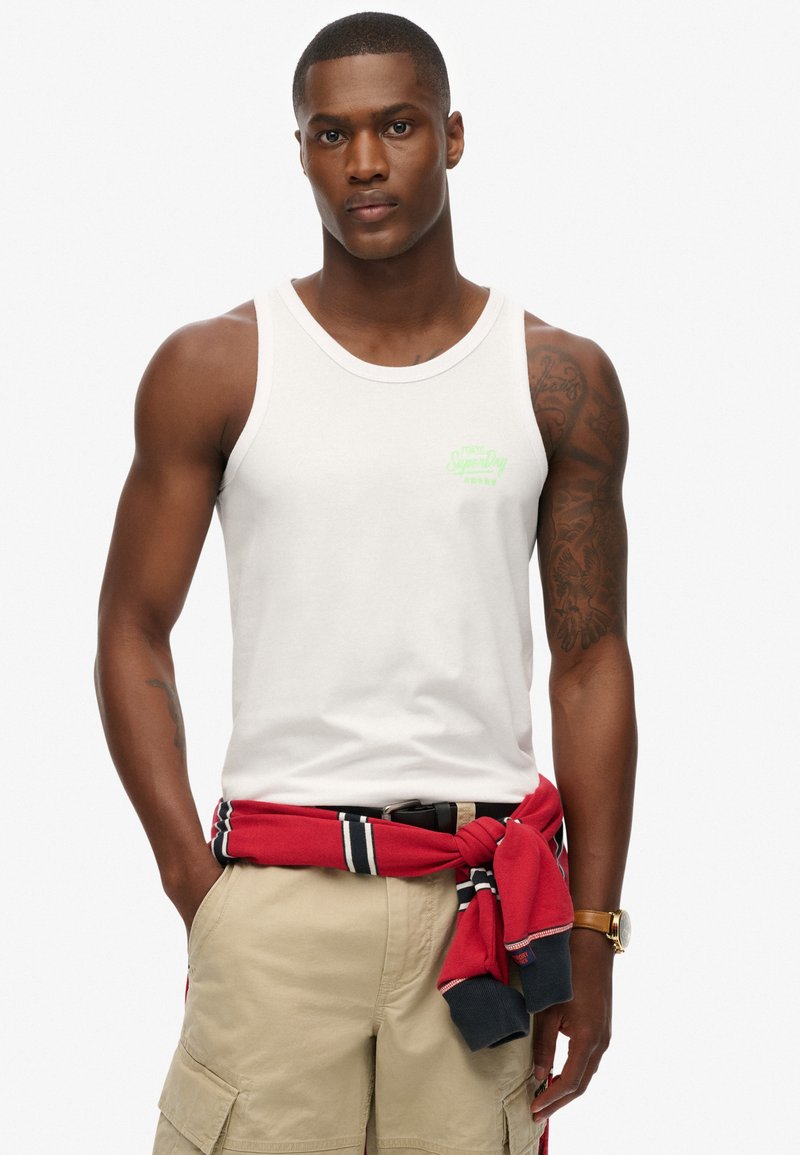 Top de tirantes de algodón blanco con logo impreso en verde, acompañado de pantalones cortos caqui y un sudadera a rayas rojas atada a la cintura.