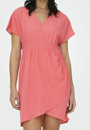 Kobieta ubrana w koralowo-różową sukienkę z krótkim rękawem, dekoltem w serek, talią podkreśloną oraz asymetrycznym dołem na białym tle.