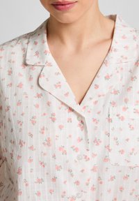 Vrouw in een witte button-up shirt met kleine roze bloemprint en een borstzakje, toont haar halslijn en kraag.