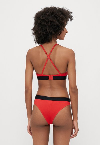 Set de lenjerie roșie format dintr-un bralette cu spate încrucișat și bretele ajustabile, împreună cu slipuri stil bikini cu un tiv negru.