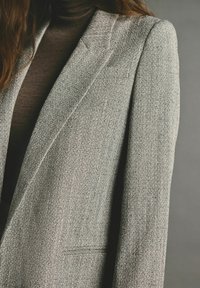 Blazer gris à motif chevron avec revers crantés, épaules structurées et texture lisse, doté d'une poche unique à l'avant.