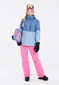 Giacca da neve impermeabile blu e azzurro, pantaloni da neve rosa, guanti neri e stivali. Tiene uno snowboard multicolore con il testo "NOW".