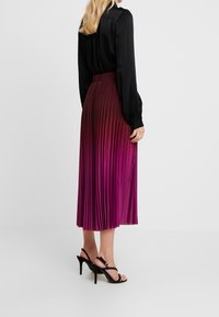 Plissierter Midirock in verlaufenden Lilatönen, fließende Textur, kombiniert mit einer schwarzen Langarmbluse und schwarzen Riemchenabsätzen.