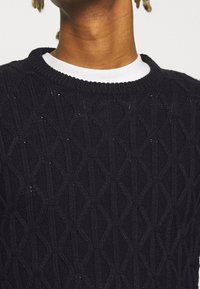 Texturerad svart stickad tröja med diamantmönster, rund hals med vit innerkrage och ribbade muddänder.