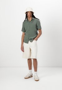 Short-sleeve green shirt, cream shorts, beige bucket hat, and light sneakers. Textured materials, relaxed fit, casual summer outfit.