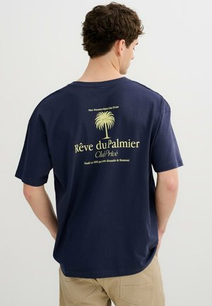 Hombre con camiseta azul marino con gráfico de palmera amarilla y texto en francés en la espalda, combinado con pantalones beige, de pie de lado.