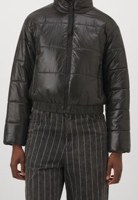 Svart vadderad jacka med glansig yta, dragkedja, hög krage och elastisk midjeresår. Har ett horisontellt quiltat mönster och sidofickor.