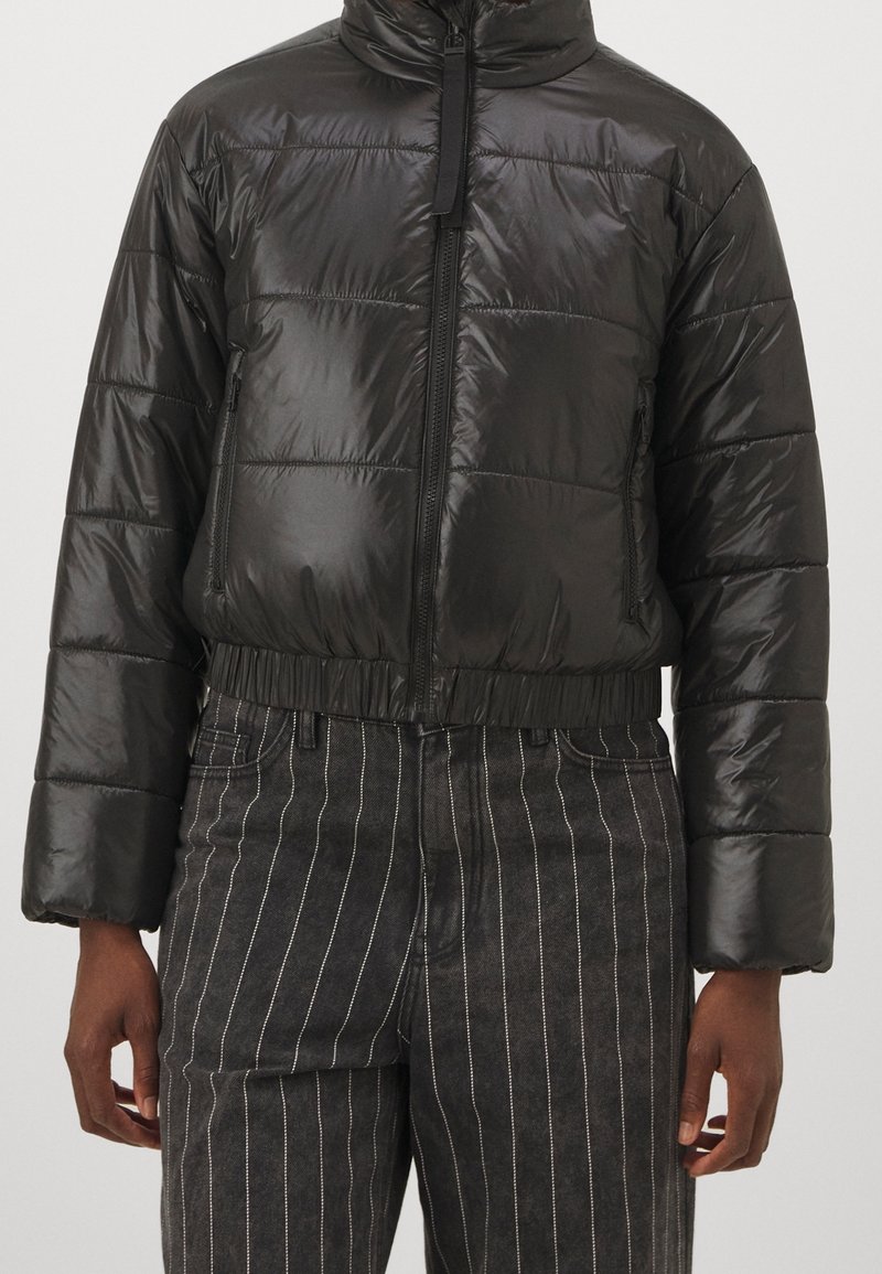 Svart vadderad jacka med glansig yta, dragkedja, hög krage och elastisk midjeresår. Har ett horisontellt quiltat mönster och sidofickor.