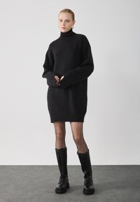 Pullover nero oversize in maglia con colletto alto, dotato di polsini e orlo a coste. Il modello indossa stivali neri con lacci fino al ginocchio.
