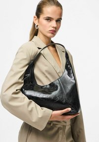 Moteris, apsirengusi kreminiu kostiumu, laiko didelę juodą blizgią rankinę su viena petneša priešais baltą foną.