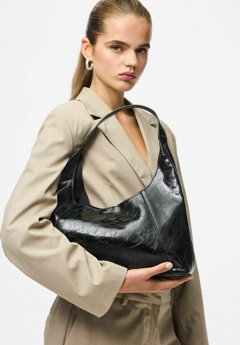 Moteris, apsirengusi kreminiu kostiumu, laiko didelę juodą blizgią rankinę su viena petneša priešais baltą foną.