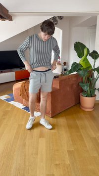 Striped gray sweater with long sleeves, and gray shorts with drawstring. White sneakers. Indoor setting with a couch and plants.