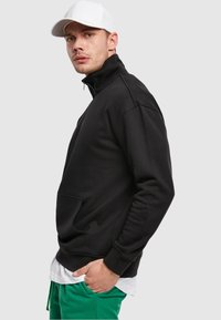 Black zip-up sweatshirt with high collar, ribbed cuffs, and front pocket. Paired with green joggers, featuring a white undershirt and cap.