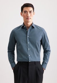 Camicia a maniche lunghe con bottone in blu navy con motivo circolare; tessuto in cotone; colletto a punta; bottoni bianchi; abbinata a pantaloni neri.