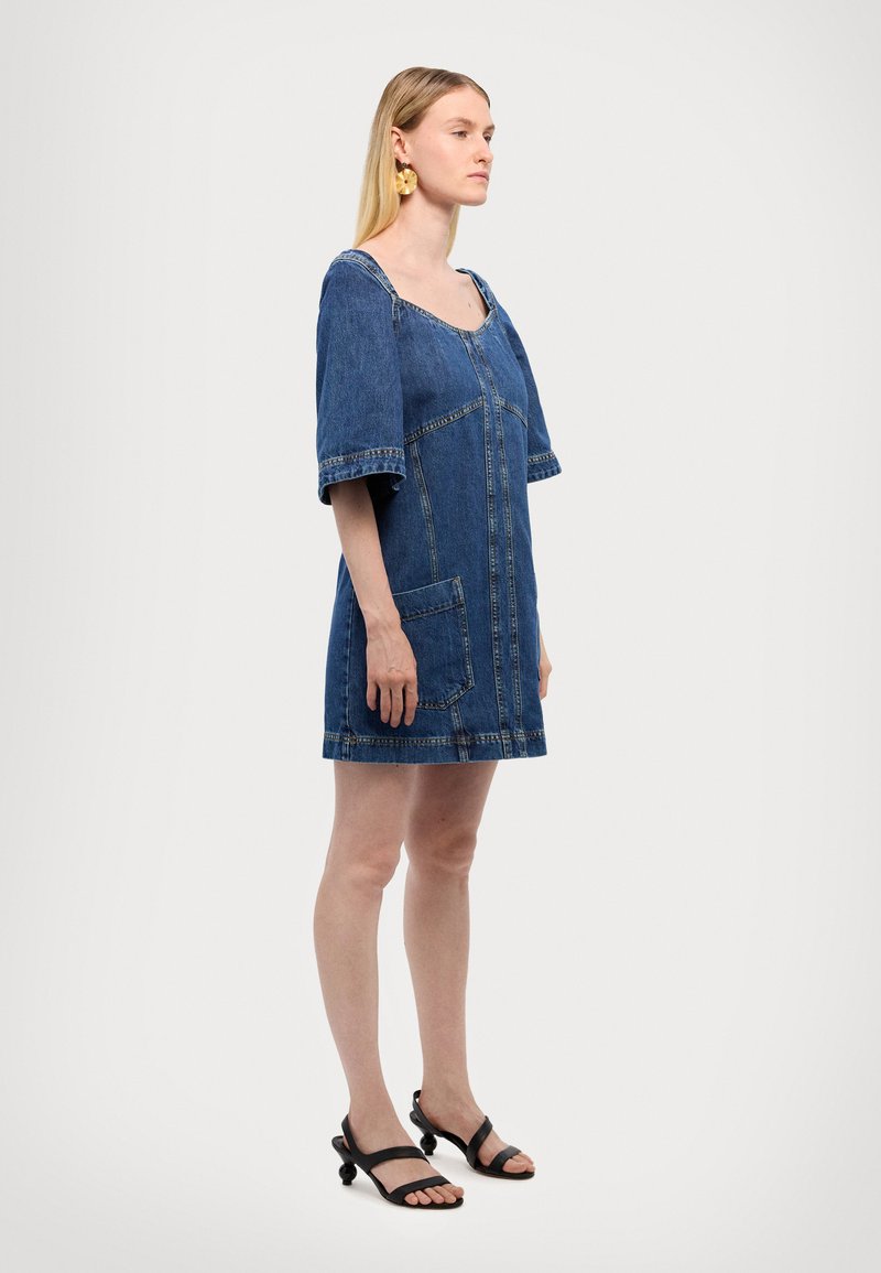 Denim obleka s kratkimi rokavi, kvadratnim izrezom, sprednjim žepom in vidnimi šivi. Nosa z črnimi sandali z okroglimi poudarki.