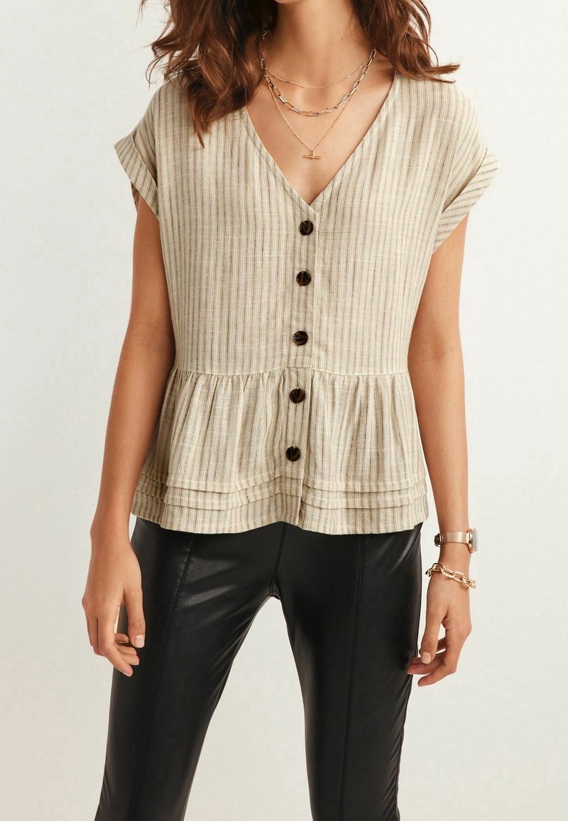 Femme portant un top beige rayé sans manches boutonné avec un ourlet volanté et un pantalon en cuir noir, accessoirisée avec des colliers dorés et des bijoux de poignet.