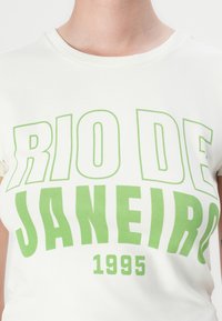 Hvit bomull t-skjorte med fet grønn tekst "RIO DE JANEIRO" og året "1995" i en sporty skrifttype. Rund hals, korte ermer.