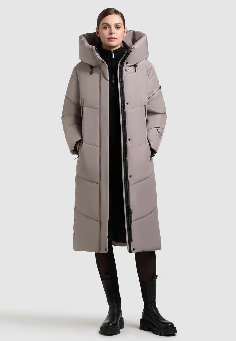 Lang, polstret frakke i lys grå med stor hætte, der har lynlås foran og trykknapper, teksturerede paneler og sidelommer.