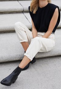 Top negro sin mangas con hombreras, pantalones crema con dobladillo enrollado y botas negras hasta el tobillo con un acabado texturizado, en un fondo de piedra.