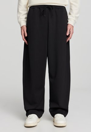 Pantaloni della tuta neri con vestibilità rilassata, elastico in vita con cordoncino e texture morbida. Abbinati a sneakers bianche.