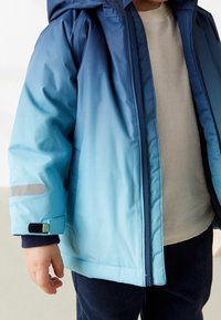 Veste à dégradé bleu et bleu clair, matelassée, avec capuche. Ses caractéristiques comprennent une fermeture éclair, des poignets élastiques et des accents réfléchissants.