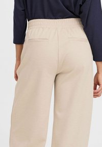 Beige byxor med elastiskt midjeband, två bakfickor, slät textur och vida ben. Tyget ser mjukt och något strukturerat ut.