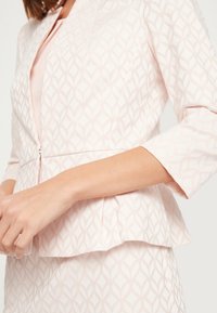 Veste rose pâle avec un motif géométrique de feuilles, design péplum et manches trois-quarts. Elle présente une fermeture éclair à l'avant et un tissu texturé.