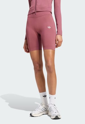 Mujer con pantalones cortos de ciclista de talle alto en color malva con logo blanco de Adidas, top a juego con cremallera, calcetines blancos Adidas y zapatillas blancas.