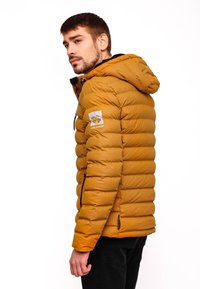 Pufová bunda v horčicovo žltej farbe, s kapucňou, horizontálnym prešívaním, dvoma zipsovými bočnými vreckami a nášivkou loga na rukáve.
