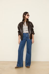Veste bomber en simili cuir marron, top court à rayures blanches et marines, jean bleu à taille haute et pattes larges, et chaussures slip-on marron.
