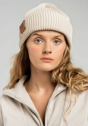 Czapka beanie w jasnym beżowym ribbowanym splocie z skórzaną metką. Model nosi dopasowaną bluzę na zamek, podkreślającą gładką teksturę i neutralny odcień.