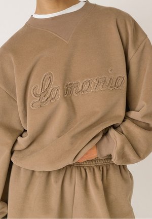 Osoba nosząca beżową, oversize bluzę i dopasowane spodnie, z napisem „La maria” wyhaftowanym kursywą na przedniej części bluzy.