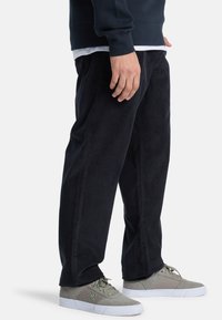 Svart kordflorsebyxor med rak bendesign, som visar strukturerad tyg och vertikala linjer. Matchas med ljusgrå sneakers.