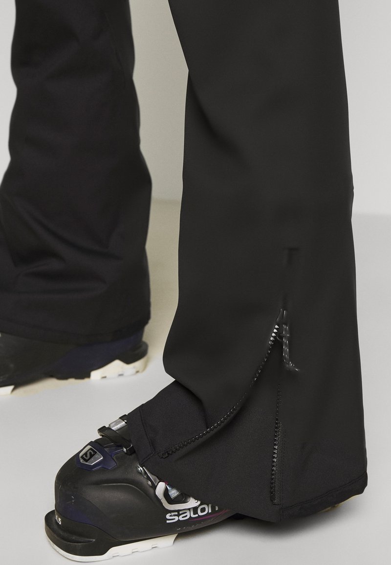 Schwarze Skihosen mit einem Reißverschluss am Saum, über Skischuhen getragen. Das Material scheint wasserabweisend und langlebig zu sein.