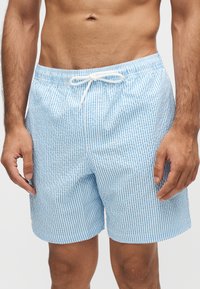 Homme portant un short de bain rayé vertical bleu et blanc avec un cordon blanc, debout, les bras détendus le long du corps.