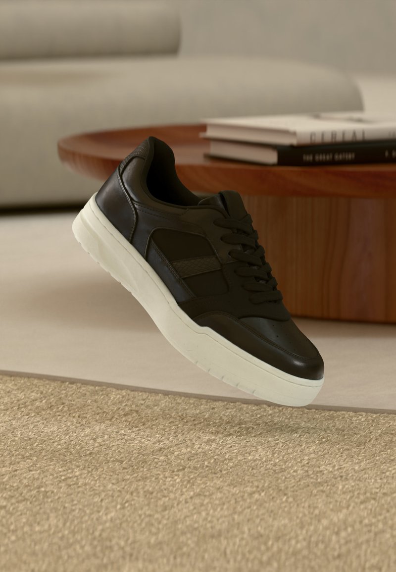 Baskets noires à lacets avec semelle blanche flottant au-dessus d'un tapis beige, avec une table en bois et des livres empilés en arrière-plan.