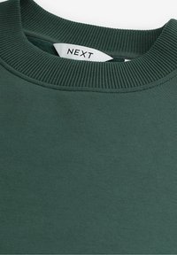 Sweat-shirt vert foncé à col rond avec col en maille côtelée et étiquette de vêtement blanche visible portant l'inscription "NEXT".