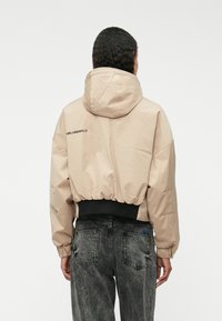 Beige rövidített dzseki kapucnival, rugalmas derékpánttal és fekete akcentusokkal. A hátulján és elülső zsebein a "KARL LAGERFELD" logó található.