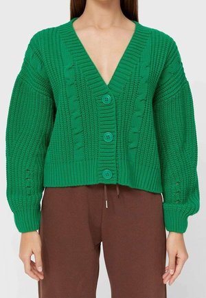 Vihreä paksu neulosta oleva neulejakku, jossa on kaapelikuvioita ja napit, puettu ruskeiden nyörivöisten housujen kanssa. Kuvassa näkyy kaulasta lantiolle.