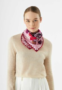 Une écharpe en soie présente un motif floral coloré sur un fond rose, avec une bordure festonnée rouge contrastante. Portée autour du cou.
