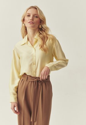 Gelbe Satinbluse mit durchgehender Knopfleiste und langen Ärmeln, kombiniert mit braunen, hoch taillierten Hosen mit einem gebundenen Taillendetail.