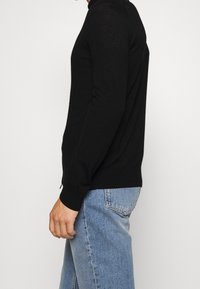 Černý svetr s dlouhým rukávem a kulatým výstřihem, vyrobený z měkké pletené látky. Přiléhavý střih s žebrovanými manžetami a lemem. Nošený s světle modrými džínami.