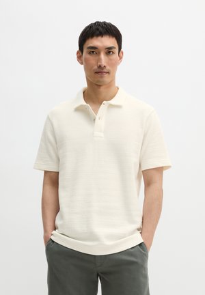 Mężczyzna z krótkimi ciemnymi włosami, ubrany w białą koszulkę polo z krótkim rękawem i ciemnoszare spodnie, stoi z rękami w kieszeniach na białym tle.